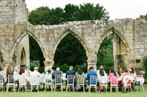 A group sit on wedding chairs outside in front of twelfth century ruins